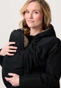 Vrouw in zwarte jas met ingebouwde babydraagzak, die een baby vasthoudt die in een bruine gebreide muts is gewikkeld, zachtjes glimlachend tegen een effen achtergrond.