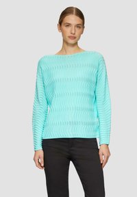 Un pull en tricot à motif vaguelé turquoise avec des manches longues et un décolleté bateau, associé à un pantalon noir. La texture est légère et respirante.