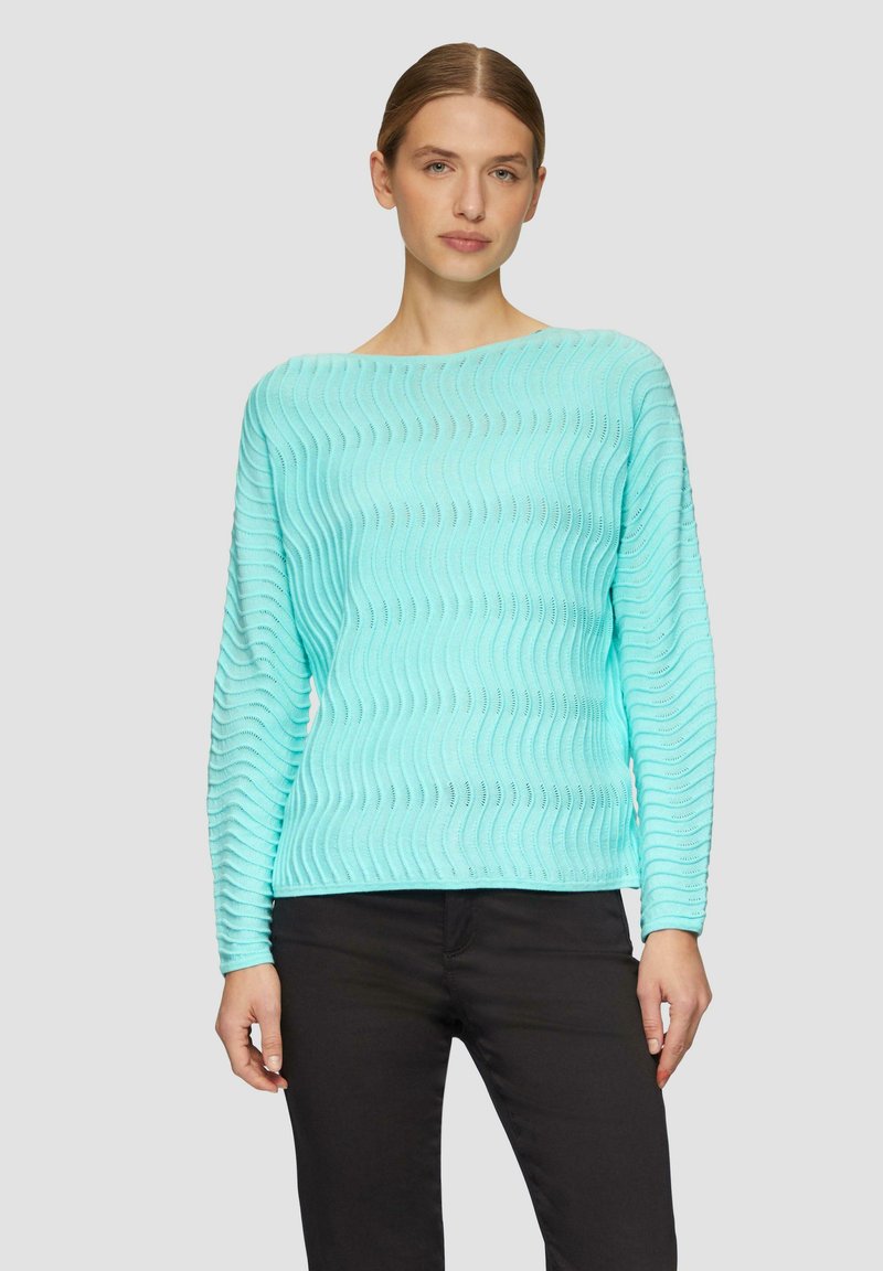 Un pull en tricot à motif vaguelé turquoise avec des manches longues et un décolleté bateau, associé à un pantalon noir. La texture est légère et respirante.