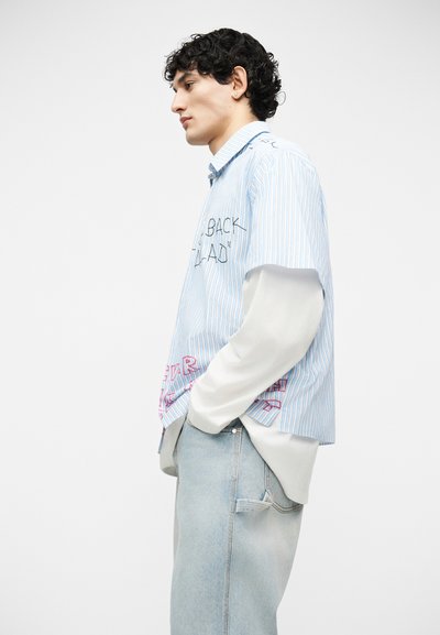 Jeune homme aux cheveux noirs bouclés portant une chemise à manches courtes à rayures bleues superposée à un haut blanc à manches longues et un jean bleu clair, les mains dans les poches.