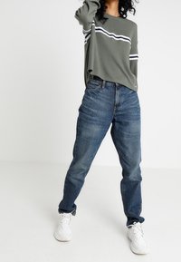 Olivgrön ribbad långärmad skjorta med vita ränder, som bärs tillsammans med högmidjade blå jeans. Vita sneakers fulländar outfiten.