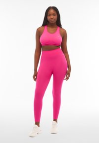Reggiseno sportivo fucsia e leggings realizzati in materiale elastico e traspirante. I leggings presentano un alto punto vita e una finitura liscia. Sneakers bianche.