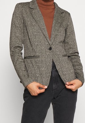 Personne portant un blazer à carreaux gris sur un col roulé marron, associé à un pantalon noir taille haute, tenant les bords du blazer avec les deux mains.