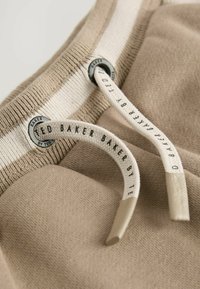Beige kangas ribaltud tekstuuriga, millel on triibuline vöökoht ja nöörid, millest igaühel on metallist tipp ja brändi detail.