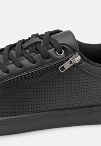 Schwarze Sneakers mit einem perforierten Obermaterial, glatter Textur, Schnürdesign und einem seitlichen Reißverschluss mit silbernen Hardwaredetails.