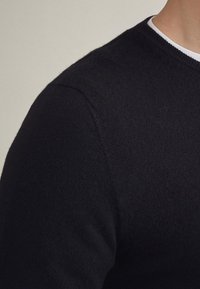 Svart stickad tröja med rund halsringning, med en mjuk textur och minimala sömmar, som visar en del av axeln och nackområdet.