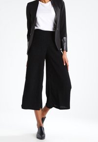 Černé široké kalhoty culottes z texturované látky, doplněné bílým tričkem a černým sako s koženými doplňky a zipy.