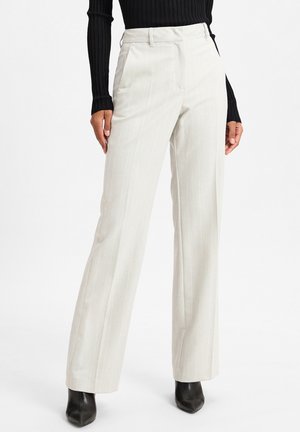 Femme portant un pantalon beige à rayures fines, taille haute et jambes larges, associé à des bottes noires à bouts pointus et un haut noir côtelé à manches longues.