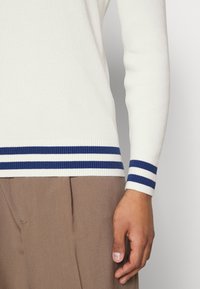 Gros plan sur une personne portant un pull blanc côtelé avec des rayures bleues au niveau des poignets et de l'ourlet, assorti à un pantalon plissé marron.