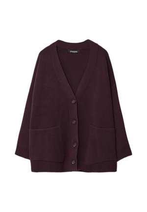 Kobieta ubrana w oversize fioletowy kardigan, biały top, szerokie ciemne dżinsy z podwiniętymi nogawkami i brązowe buty, stojąca na jednolitym tle.
