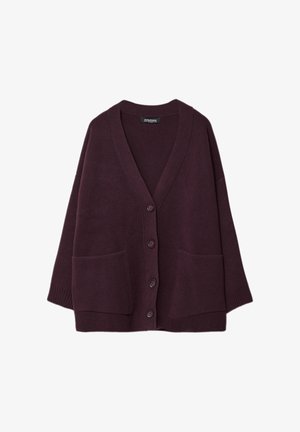 Kobieta ubrana w oversize fioletowy kardigan, biały top, szerokie ciemne dżinsy z podwiniętymi nogawkami i brązowe buty, stojąca na jednolitym tle.