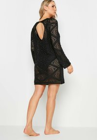 Vestido de crochet negro con mangas largas, detalle de espalda abierta, patrón de diamantes y silueta ajustada. La tela texturizada permite la visibilidad.