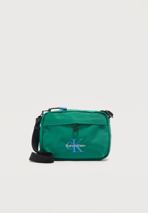 Groene compacte crossbodytas met zwarte verstelbare riem, voorvak met ritssluiting en blauw Calvin Klein-logo op witte achtergrond.