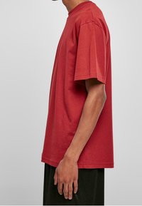 Vue de côté d'une personne portant un t-shirt rouge ample à manches courtes et un pantalon noir sur un fond gris clair uni.