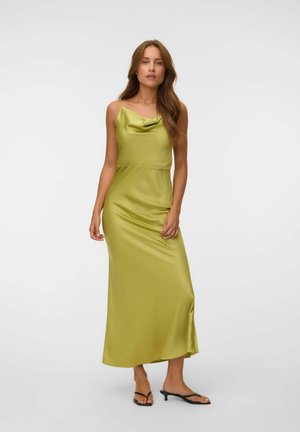 Femme debout portant une longue robe en satin vert citron sans manches avec fines bretelles et des sandales noires à talons bas sur un fond blanc uni.