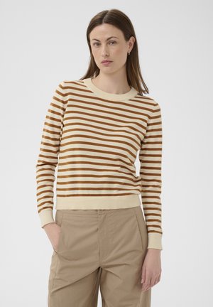 Vrouw met steil bruin haar, gekleed in een beige en bruin gestreepte trui en beige broek, staand met één hand in haar zak.