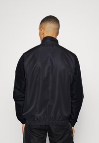Chaqueta ligera negra con un alto cuello y dobladillo elástico, que presenta una textura suave y brillante y mangas largas. Se muestra la vista posterior.