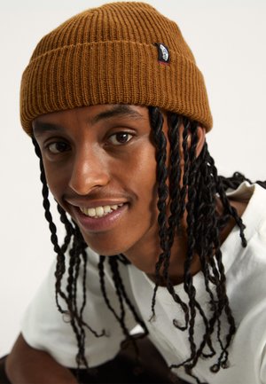 Brown ribbed knit beanie with a snug fit, featuring a small logo tag. The wearer has long, twisted hair and is dressed in a white shirt.