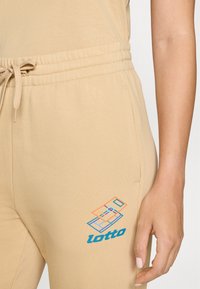Bežinės sportinės kelnės su elastingu juosmeniu, dekoratyviu raišteliu ir daugiak aspiriniu „lotto“ logotipu ant šlaunies. Minkšta, glotni tekstūra.