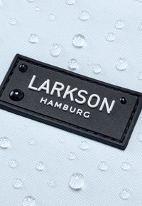Svetlomodrá tkanina s vodnými kvapkami, obsahujúca čierny obdĺžnikový štítok s embosovaným textom "LARKSON HAMBURG" bielymi písmenami a zaoblenými akcentmi.