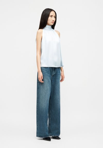 Kvinne med langt mørkt hår iført ermelløs lyseblå bluse med høy hals, vide blå jeans og svarte spisse sko, står vendt mot høyre.