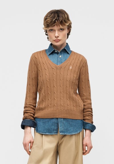 Personne portant un pull marron à col en V en maille torsadée superposé à une chemise en denim bleu et un pantalon beige, debout avec les mains détendues.