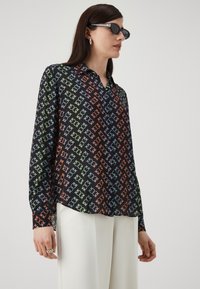 Crna bluza s geometrijskim uzorkom u narančastoj, zelenoj i plavoj boji. Dugi rukavi, kragna s dugmadi i glatka tkanina. Uparena s bijelim hlačama.