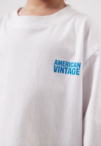 Gros plan sur une chemise blanche avec le texte bleu "AMERICAN VINTAGE" imprimé sur la partie supérieure gauche de la poitrine.