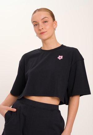 T-shirt nera cropped in cotone con scollo rotondo e maniche larghe, caratterizzata da un piccolo ricamo floreale rosa sul petto.