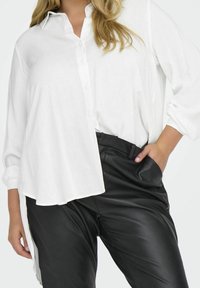 Camicia bianca button-up con maniche lunghe, vestibilità rilassata e spacchi laterali, abbinata a pantaloni in pelle sintetica nera. Tessuto leggero e morbido.