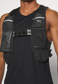 Veste de course noire avec plusieurs poches, tissu en maille, bretelles réglables et accents réfléchissants. Le devant comprend un porte-identification transparent.