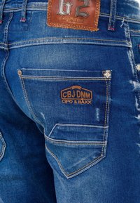Blauwe denim jeans hebben een leren label met de tekst "CIPO & BAXX" en stikseldetails. De achterzak bevat een geborduurd logo.