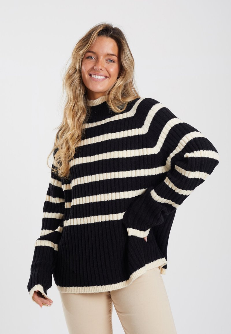 Pull en tricot rayé noir et crème avec texture côtelée, col haut et épaules tombantes. Comprend des fentes sur les côtés et des manches longues.