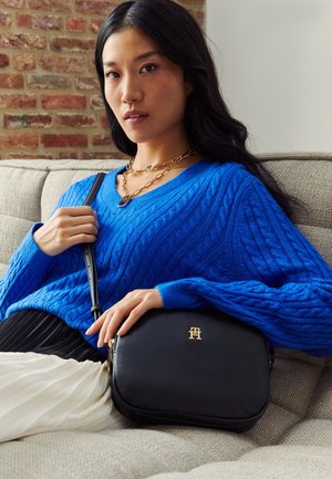 Woman with long black hair wearing a blue cable-knit sweater, gold chain necklace, and holding a black handbag with gold logo while seated on a beige couch.