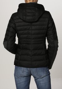 Sort polstret jakke med hætte, med kviltet tekstur, grå accenter på ærmerne og en tætsiddende pasform. Bæres over blå jeans.