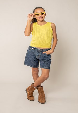 Fille portant un haut jaune sans manches, un short en jean, des bottes marron et des lunettes de soleil jaunes, souriant avec une main sur la hanche et l'autre ajustant ses lunettes de soleil.