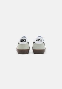 Zapatillas Nike en blanco y gris que presentan una parte trasera de ante, acentos negros, suela de goma y un gran logotipo "NIKE" en el talón.