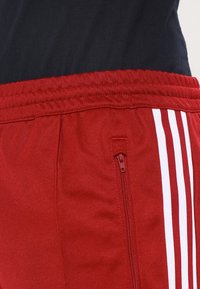 Červené sportovní šortky s texturovanou tkaninou, elastickým pasem a boční kapsou na zip. Na jedné straně jsou tři bílé pruhy.