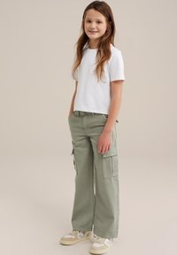 Balta īsām piedurknēm T-kreklu, ko apvieno ar gaiši zaļām kravas biksēm, kurām ir sānu kabatas. Valkā ar bēšiem sporta apaviem, kuros ir baltas akcenti.