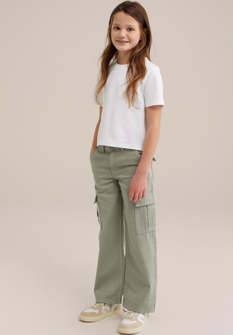 Balta īsām piedurknēm T-kreklu, ko apvieno ar gaiši zaļām kravas biksēm, kurām ir sānu kabatas. Valkā ar bēšiem sporta apaviem, kuros ir baltas akcenti.