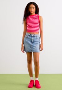 Top corto a righe rosse e bianche, gonna di jeans azzurri chiari, sneakers rosa e calzini bianchi, in piedi su un pavimento verde con uno sfondo semplice.