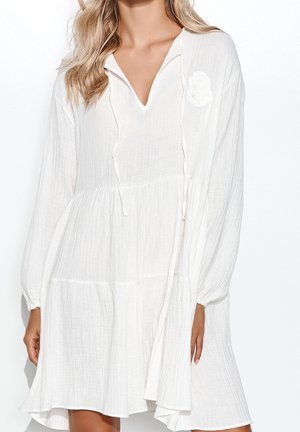 Femme portant une robe blanche texturée à manches longues avec un col en V et une jupe à volants, avec un petit emblème brodé sur la poitrine.