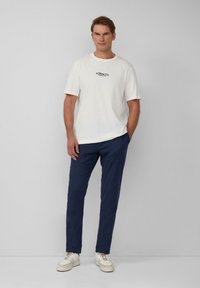 Witte t-shirt met korte mouwen en een marineblauw logo, gedragen met marineblauwe broek en witte sneakers met beige accenten. Eenvoudig, strak ontwerp.