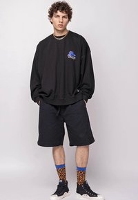Sudadera negra de gran tamaño con gráfico azul, combinada con pantalones cortos negros. Los accesorios incluyen calcetines de estampado de leopardo y zapatillas altas negras.