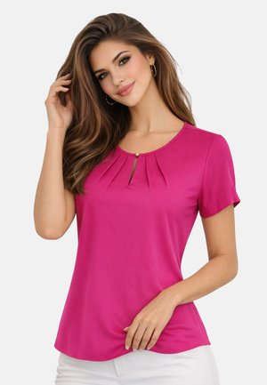 Kobieta o długich brązowych włosach, ubrana w różową bluzkę z krótkim rękawem z plisowanym dekoltem i białe spodnie, dotyka włosów i się uśmiecha.