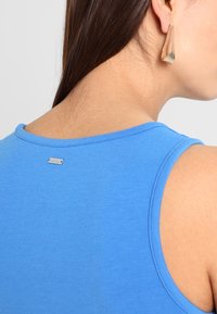 Niebieski top bez rękawów wykonany z miękkiego materiału, z okrągłym dekoltem i małym srebrnym logo z tyłu. Gładka tekstura i dopasowany krój.