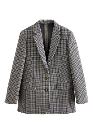 Frau lächelnd, trägt schwarze Bluse, weite schwarze Hose, karierten Blazer, braunen Gürtel, mit Händen in den Taschen auf neutralem Hintergrund.