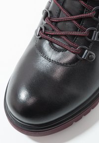 Botte en cuir noir à bout rond, lacets bordeaux et noirs passés dans des œillets métalliques, semelle épaisse bordeaux sur fond blanc.