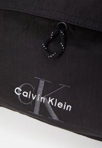 Zwarte stoffen tas met een trekkoordsluiting. Geborduurd "Calvin Klein" logo in wit en grijs. Gestructureerd oppervlak en minimalistisch ontwerp.