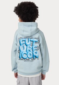 Felpa azzurra chiara con una grande scritta stilizzata "FUTURE ICON" in blu, carattere testurizzato sul retro, circondata da testo nero e grafiche di faccine sorridenti.